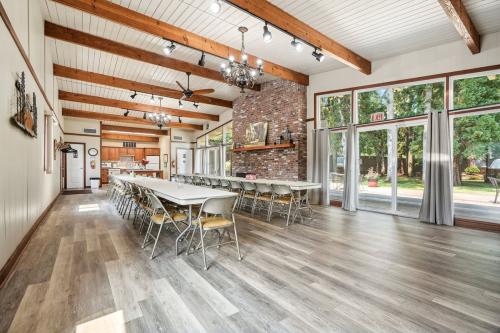 A large, well-lit room with exposed wooden beams, a brick fireplace, and long tables with chairs set up for a meeting or event. The room has large windows and wood flooring.