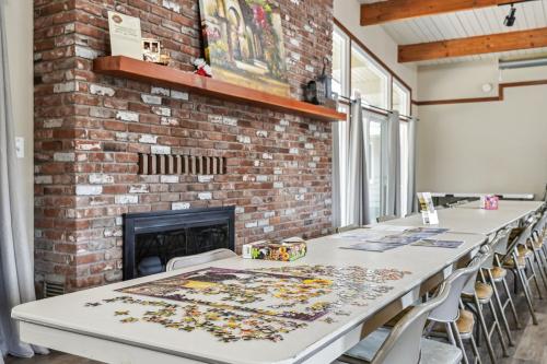 A long table partially covered with a jigsaw puzzle sits in front of a brick fireplace in a room with exposed wooden beams and large windows.
