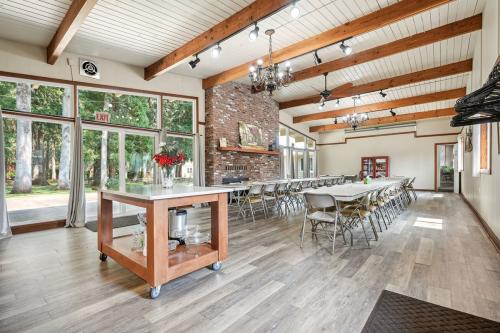 A spacious, well-lit room with long tables and chairs set up for a gathering. The room features wooden beams on the ceiling, a brick fireplace, and large windows overlooking a grassy outdoor area.