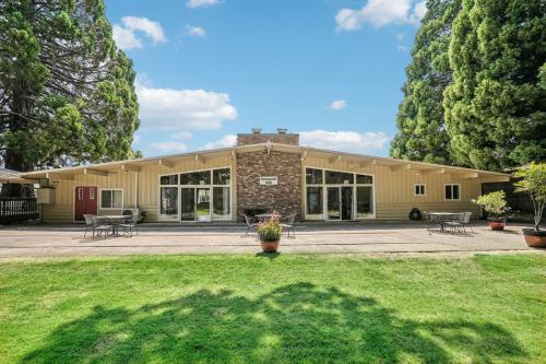 A single-story building with large windows and a stone facade sits amidst tall trees and a grassy area under a clear blue sky.