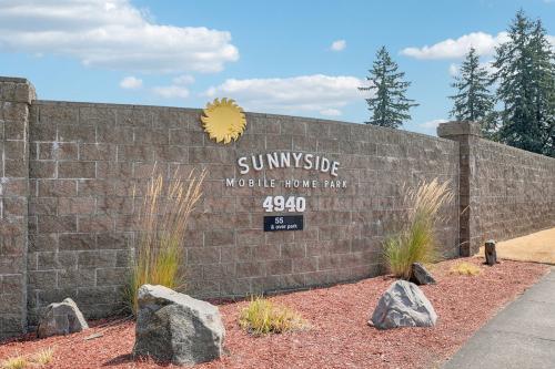 Entrance to Sunnyside Mobile Home Park showing a stone wall with the park's name, address number (4940), and a sun emblem, surrounded by landscaping with rocks and grass.