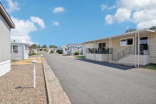 A paved road runs between rows of mobile homes in a residential area. The sky is clear with a few clouds, and small shrubs are visible along the street.