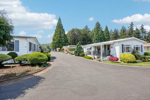 A quiet residential street lined with white mobile homes, manicured lawns, and green trees under a blue sky with scattered clouds.